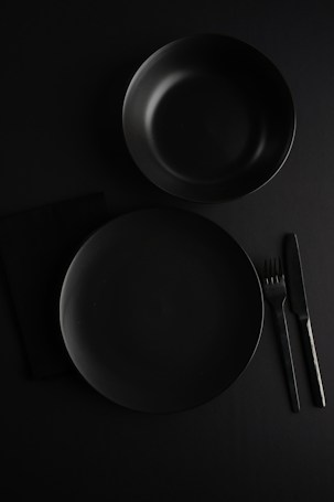 A minimalistic flat lay setup featuring a dark-colored dinner plate and bowl. Positioned on a black surface, the arrangement includes a knife and fork to the right of the plate. The overall composition emphasizes simplicity and elegance, with a focus on clear lines and shapes.