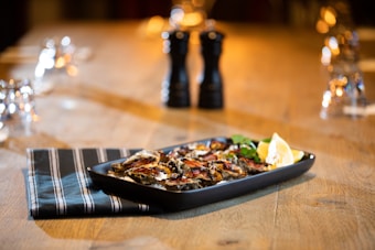 A wooden dining table is set with a black rectangular plate containing a grilled dish, possibly eggplant or mushrooms, garnished with lemon wedges and herbs. Two pepper grinders stand in the background, surrounded by reflections from glassware, creating a warm, inviting atmosphere.