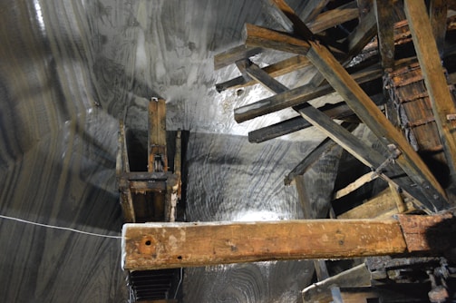 A complex structure made of wooden beams and planks is positioned against a textured surface that appears to be a rock or cave wall. The surfaces seem damp, reflecting light in certain areas, and the beams intersect at various angles, suggesting part of an old industrial or mining setup.