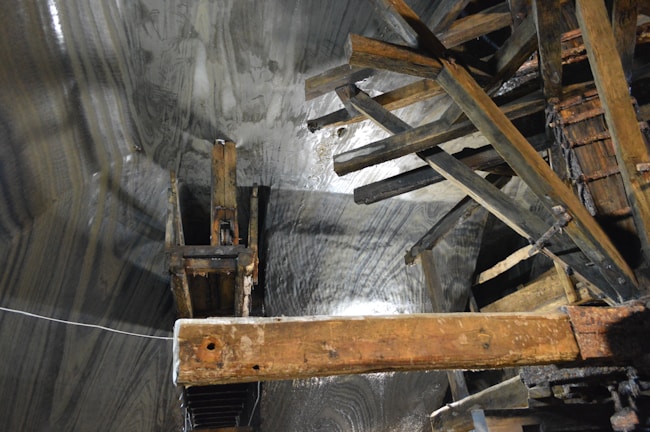 A complex structure made of wooden beams and planks is positioned against a textured surface that appears to be a rock or cave wall. The surfaces seem damp, reflecting light in certain areas, and the beams intersect at various angles, suggesting part of an old industrial or mining setup.