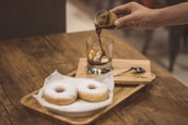 A hand is pouring espresso over a scoop of ice cream in a glass, creating an affogato. Two powdered sugar donuts are placed on a wooden tray nearby. The scene is set on a wooden table, conveying a cozy coffee shop atmosphere.