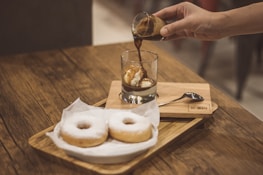 A hand is pouring espresso over a scoop of ice cream in a glass, creating an affogato. Two powdered sugar donuts are placed on a wooden tray nearby. The scene is set on a wooden table, conveying a cozy coffee shop atmosphere.