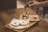 A hand is pouring espresso over a scoop of ice cream in a glass, creating an affogato. Two powdered sugar donuts are placed on a wooden tray nearby. The scene is set on a wooden table, conveying a cozy coffee shop atmosphere.