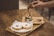 A hand is pouring espresso over a scoop of ice cream in a glass, creating an affogato. Two powdered sugar donuts are placed on a wooden tray nearby. The scene is set on a wooden table, conveying a cozy coffee shop atmosphere.