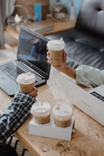 Two laptops on a wooden table with several paper coffee cups featuring a green logo. Two hands are seen holding some of the cups, indicating a shared coffee moment. The background includes a blurred shelf with books and a decorative item.