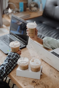Two laptops on a wooden table with several paper coffee cups featuring a green logo. Two hands are seen holding some of the cups, indicating a shared coffee moment. The background includes a blurred shelf with books and a decorative item.