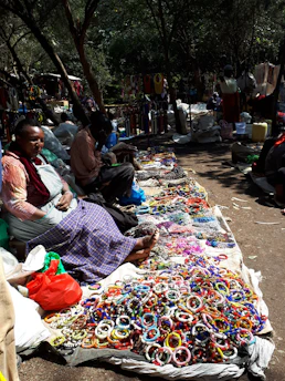 A lively outdoor market setting with vendors sitting on the ground displaying a colorful array of handmade bracelets. The scene is shaded by trees, and various shoppers are visible in the background, engaged in browsing and purchasing items. The ground is covered with woven mats on which the jewelry is neatly arranged, and some bags and containers are scattered around.