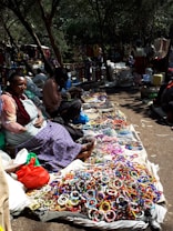 A lively outdoor market setting with vendors sitting on the ground displaying a colorful array of handmade bracelets. The scene is shaded by trees, and various shoppers are visible in the background, engaged in browsing and purchasing items. The ground is covered with woven mats on which the jewelry is neatly arranged, and some bags and containers are scattered around.