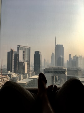 Silhouette of a person's feet resting by a large window with a view of a city skyline featuring modern skyscrapers and the iconic Burj Khalifa. The scene is bathed in a hazy, ambient light, suggesting a sunrise or sunset.