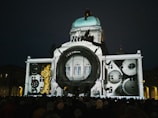 An architectural building is illuminated with a projection of clock gears and camera lens elements. The scene is set at night with a crowd of people gathered in front of the building.