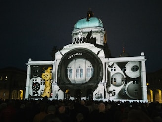 An architectural building is illuminated with a projection of clock gears and camera lens elements. The scene is set at night with a crowd of people gathered in front of the building.