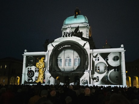 An architectural building is illuminated with a projection of clock gears and camera lens elements. The scene is set at night with a crowd of people gathered in front of the building.