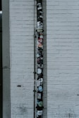 A vertical drainpipe is affixed to a white brick wall and adorned with various stickers. The wall is slightly weathered, with visible lines of horizontal bricks. The pipe, which appears black, is covered in colorful stickers that include various shapes, logos, and text, adding a vibrant contrast to the otherwise monochromatic background.