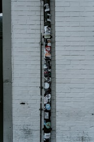 A vertical drainpipe is affixed to a white brick wall and adorned with various stickers. The wall is slightly weathered, with visible lines of horizontal bricks. The pipe, which appears black, is covered in colorful stickers that include various shapes, logos, and text, adding a vibrant contrast to the otherwise monochromatic background.