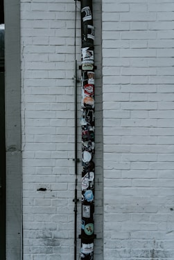 A vertical drainpipe is affixed to a white brick wall and adorned with various stickers. The wall is slightly weathered, with visible lines of horizontal bricks. The pipe, which appears black, is covered in colorful stickers that include various shapes, logos, and text, adding a vibrant contrast to the otherwise monochromatic background.