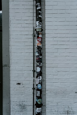 A vertical drainpipe is affixed to a white brick wall and adorned with various stickers. The wall is slightly weathered, with visible lines of horizontal bricks. The pipe, which appears black, is covered in colorful stickers that include various shapes, logos, and text, adding a vibrant contrast to the otherwise monochromatic background.
