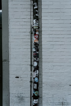 A vertical drainpipe is affixed to a white brick wall and adorned with various stickers. The wall is slightly weathered, with visible lines of horizontal bricks. The pipe, which appears black, is covered in colorful stickers that include various shapes, logos, and text, adding a vibrant contrast to the otherwise monochromatic background.