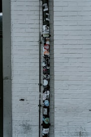 A vertical drainpipe is affixed to a white brick wall and adorned with various stickers. The wall is slightly weathered, with visible lines of horizontal bricks. The pipe, which appears black, is covered in colorful stickers that include various shapes, logos, and text, adding a vibrant contrast to the otherwise monochromatic background.