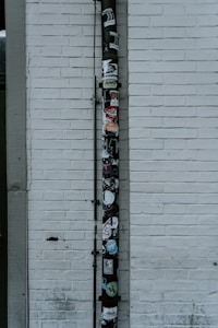 A vertical drainpipe is affixed to a white brick wall and adorned with various stickers. The wall is slightly weathered, with visible lines of horizontal bricks. The pipe, which appears black, is covered in colorful stickers that include various shapes, logos, and text, adding a vibrant contrast to the otherwise monochromatic background.