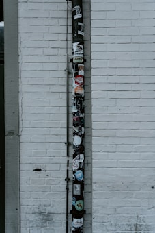 A vertical drainpipe is affixed to a white brick wall and adorned with various stickers. The wall is slightly weathered, with visible lines of horizontal bricks. The pipe, which appears black, is covered in colorful stickers that include various shapes, logos, and text, adding a vibrant contrast to the otherwise monochromatic background.
