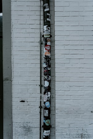 A vertical drainpipe is affixed to a white brick wall and adorned with various stickers. The wall is slightly weathered, with visible lines of horizontal bricks. The pipe, which appears black, is covered in colorful stickers that include various shapes, logos, and text, adding a vibrant contrast to the otherwise monochromatic background.