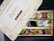 An open box of assorted traditional Indian sweets, known as mithai, with a variety of bright and colorful pieces arranged neatly in compartments. The box displays elegant gold floral designs and inscriptions, with the label 'Dryfruit Sweets Premium 24 Carats Mithai Magic' on the lid.