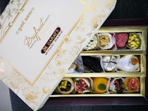 An open box of assorted traditional Indian sweets, known as mithai, with a variety of bright and colorful pieces arranged neatly in compartments. The box displays elegant gold floral designs and inscriptions, with the label 'Dryfruit Sweets Premium 24 Carats Mithai Magic' on the lid.