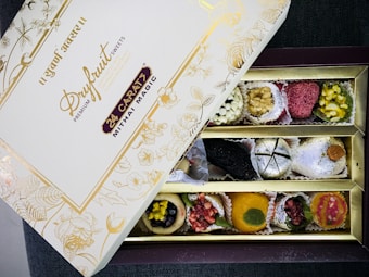 An open box of assorted traditional Indian sweets, known as mithai, with a variety of bright and colorful pieces arranged neatly in compartments. The box displays elegant gold floral designs and inscriptions, with the label 'Dryfruit Sweets Premium 24 Carats Mithai Magic' on the lid.