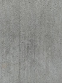 A close-up view of a textured concrete surface, featuring a noticeable crack running vertically. The surface displays subtle variations in shading and slight discolorations, with small speckles and irregularities visible across the surface.