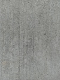 A close-up view of a textured concrete surface, featuring a noticeable crack running vertically. The surface displays subtle variations in shading and slight discolorations, with small speckles and irregularities visible across the surface.