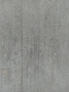 A close-up view of a textured concrete surface, featuring a noticeable crack running vertically. The surface displays subtle variations in shading and slight discolorations, with small speckles and irregularities visible across the surface.