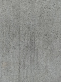 A close-up view of a textured concrete surface, featuring a noticeable crack running vertically. The surface displays subtle variations in shading and slight discolorations, with small speckles and irregularities visible across the surface.