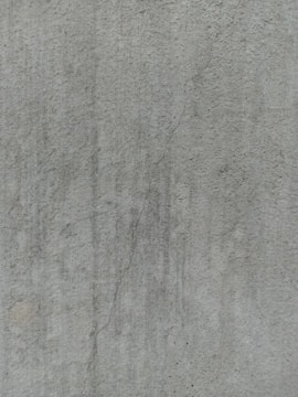 A close-up view of a textured concrete surface, featuring a noticeable crack running vertically. The surface displays subtle variations in shading and slight discolorations, with small speckles and irregularities visible across the surface.