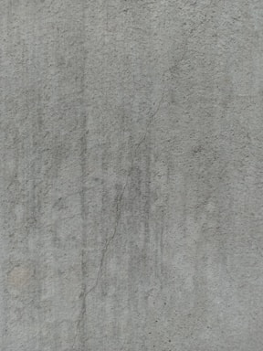 A close-up view of a textured concrete surface, featuring a noticeable crack running vertically. The surface displays subtle variations in shading and slight discolorations, with small speckles and irregularities visible across the surface.