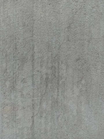 A close-up view of a textured concrete surface, featuring a noticeable crack running vertically. The surface displays subtle variations in shading and slight discolorations, with small speckles and irregularities visible across the surface.