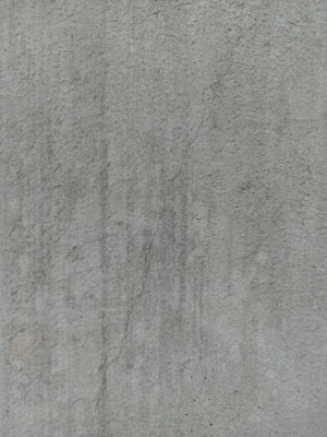 A close-up view of a textured concrete surface, featuring a noticeable crack running vertically. The surface displays subtle variations in shading and slight discolorations, with small speckles and irregularities visible across the surface.