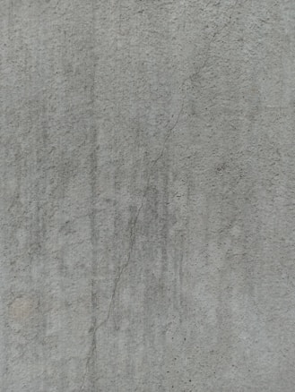 A close-up view of a textured concrete surface, featuring a noticeable crack running vertically. The surface displays subtle variations in shading and slight discolorations, with small speckles and irregularities visible across the surface.