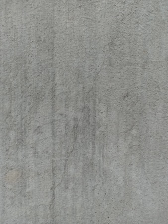 A close-up view of a textured concrete surface, featuring a noticeable crack running vertically. The surface displays subtle variations in shading and slight discolorations, with small speckles and irregularities visible across the surface.