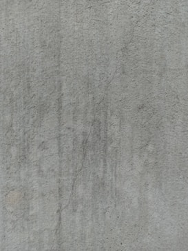 A close-up view of a textured concrete surface, featuring a noticeable crack running vertically. The surface displays subtle variations in shading and slight discolorations, with small speckles and irregularities visible across the surface.