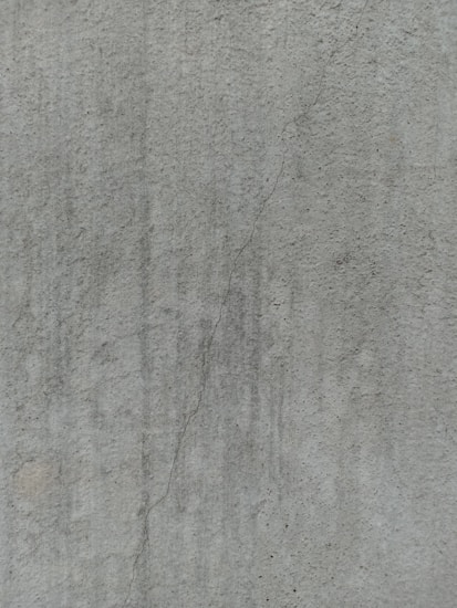 A close-up view of a textured concrete surface, featuring a noticeable crack running vertically. The surface displays subtle variations in shading and slight discolorations, with small speckles and irregularities visible across the surface.