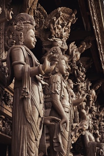 Intricately carved wooden statues with detailed, ornate features depicting human-like figures adorned in traditional attire. The sculptures present a sense of grandeur and artistic craftsmanship, showcasing detailed facial expressions and elaborate headdresses.
