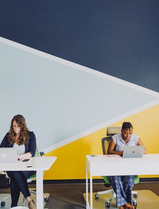 two women sitting beside table using MacBooks