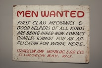 A vintage sign with bold red and black lettering on a white background. The text advertises job openings for first-class mechanics and helpers, with instructions to contact Charles Schmidt for an application. The bottom features the name of a shipbuilding and dry dock company located in Sturgeon Bay, Wisconsin.