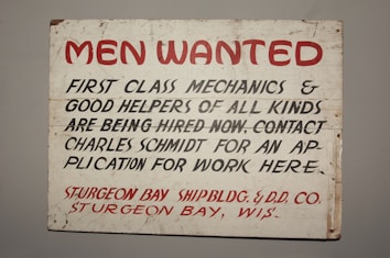 A vintage sign with bold red and black lettering on a white background. The text advertises job openings for first-class mechanics and helpers, with instructions to contact Charles Schmidt for an application. The bottom features the name of a shipbuilding and dry dock company located in Sturgeon Bay, Wisconsin.