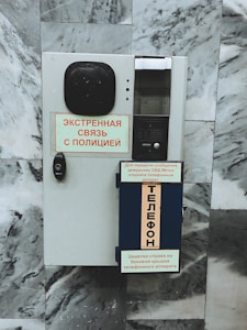 A wall-mounted emergency communication device with text in Russian. It includes a speaker, buttons, and instruction signs. The background consists of textured gray marble tiles.