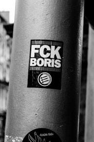 A black and white photograph of a circular metal pole with a sticker that expresses a political message. The sticker features bold white letters on a black background and includes an anti-fascism logo.