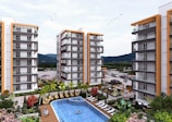 A modern residential complex featuring multiple high-rise buildings with balconies. In the center, there is a swimming pool surrounded by lounge chairs and landscaping with trees and colorful flowers. Cars are parked in the distance, and the area is set against a backdrop of mountains under a partly cloudy sky with birds flying overhead.