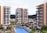 A modern residential complex featuring multiple high-rise buildings with balconies. In the center, there is a swimming pool surrounded by lounge chairs and landscaping with trees and colorful flowers. Cars are parked in the distance, and the area is set against a backdrop of mountains under a partly cloudy sky with birds flying overhead.