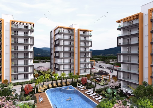 A modern residential complex featuring multiple high-rise buildings with balconies. In the center, there is a swimming pool surrounded by lounge chairs and landscaping with trees and colorful flowers. Cars are parked in the distance, and the area is set against a backdrop of mountains under a partly cloudy sky with birds flying overhead.