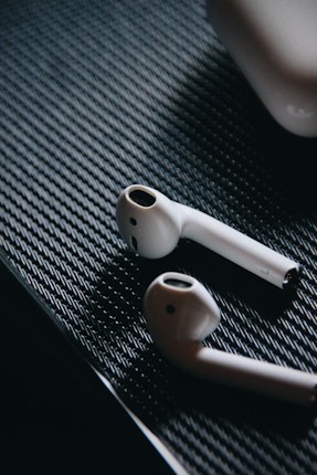 White wireless earbuds are placed on a textured black surface, with an accompanying charging case visible in the top right corner. The lighting casts subtle shadows, emphasizing the smooth and sleek design of the earbuds.